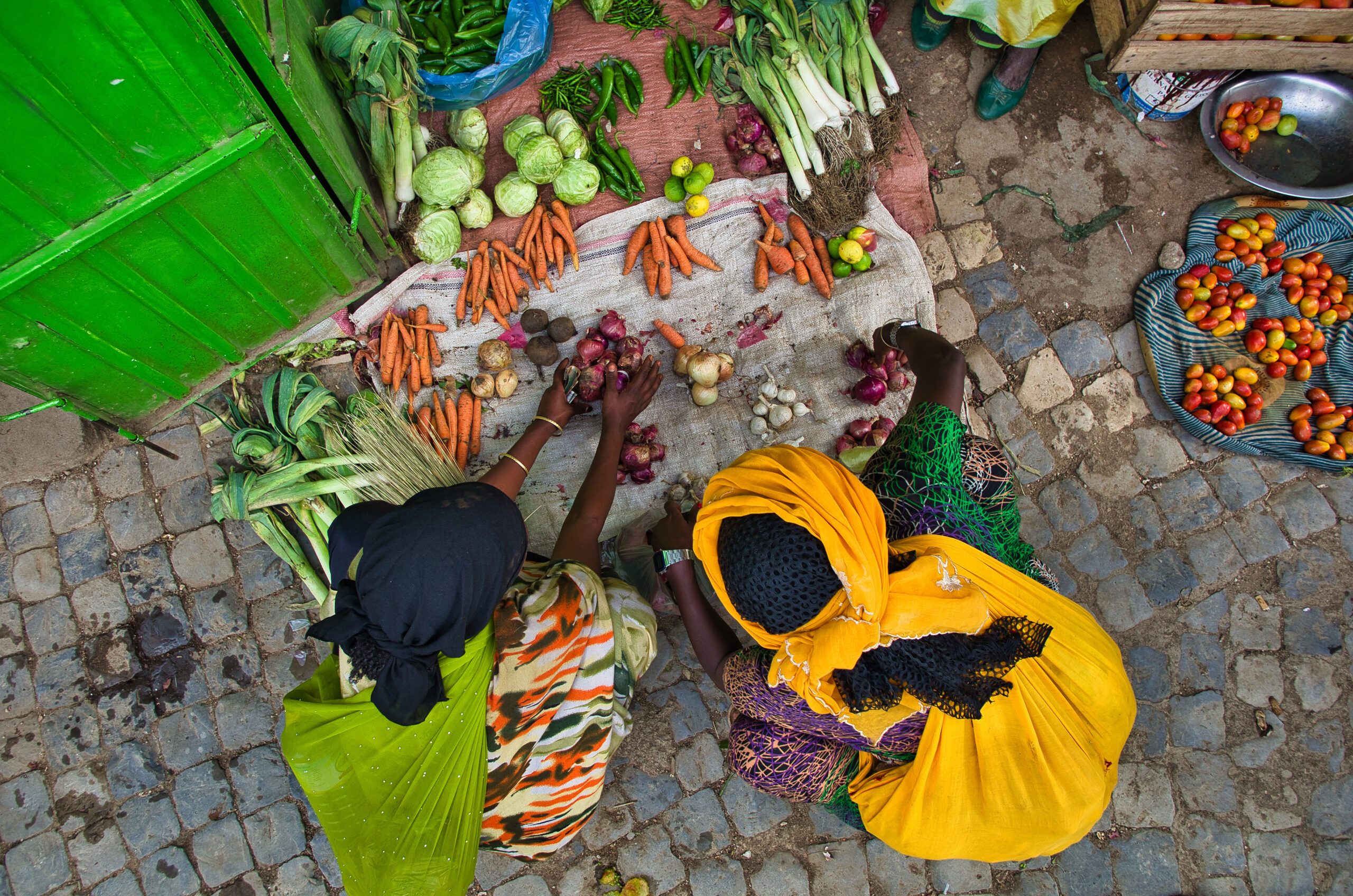 Black women in colorful traditional African costumes crouching at a vegetable stand on the ground and buying vegetables. Take a zenith. Carrots, tomatoes, onions, garlic, leeks, peppers. Women in colorful traditional African costumes crouching at a vegetable stand on the ground and buying vegetables. Take a zenith. Carrots, tomatoes, onions, garlic, leeks, peppers.